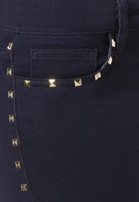Jeans de mezclilla azul oscuro con tachuelas en forma de pirámide doradas a lo largo de la costura lateral y el bolsillo. Textura suave con un diseño ajustado.