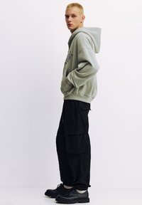 Hanorac gri deschis cu glugă și buzunar frontal, asortat cu pantaloni cargo negri largi cu buzunare laterale și pantofi negri cu șireturi.