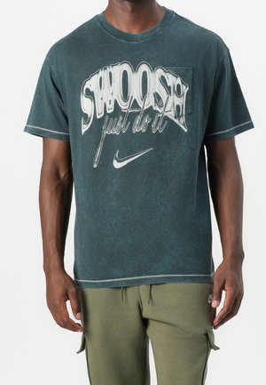 Mężczyzna w ciemnozielonej koszulce z białym napisem "SWOOSH just do it" i logo Nike, w połączeniu z oliwkowymi spodniami typu cargo.