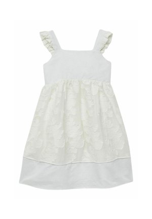 Młoda dziewczyna w białej sukience bez rękawów z kwiatowym wzorem i złotych butach Mary Jane, stoi z jedną nogą lekko ugiętą.