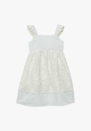 Niña joven con vestido blanco sin mangas con textura floral y zapatos Mary Jane dorados, de pie con una pierna ligeramente doblada.