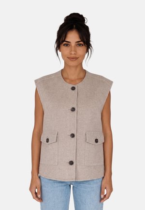 Femme aux cheveux foncés attachés, portant un gilet beige sans manches boutonné avec des poches devant et un jean bleu clair, se tenant devant un fond uni.