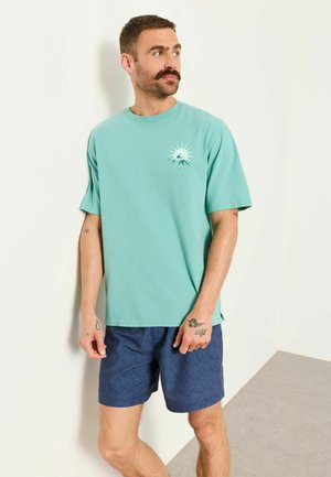 Mann mit Schnurrbart und Tattoos, der ein türkisfarbenes Grafik-T-Shirt und marineblaue gemusterte Shorts trägt, steht vor einer weißen Wand und schaut nach rechts.