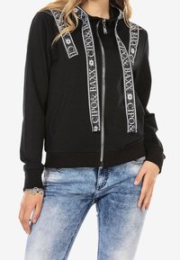 Black zip-up jacket with white patterned stripes, featuring a stand collar, long sleeves, and front pockets. Paired with blue denim jeans.