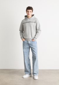Grå hettegenser med en svart logo over brystet, sammen med lyseblå vide jeans og hvite sneakers, stående mot en nøytral bakgrunn.