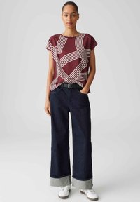 Maronfarbener, gemusterter Oberteil mit weißen Streifen, kombiniert mit weiten, dunklen Jeans mit umgeschlagenem grauen Saum. Weiße Sneaker runden den Look ab.