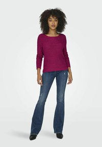 Pull en tricot magenta à manches longues, col rond, associé à un jean évasé bleu foncé. Texture lisse, motif subtil et coupe décontractée.