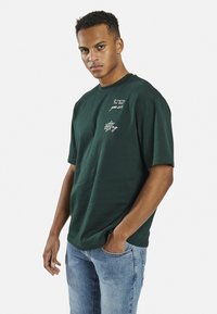 Grünes Baumwoll-T-Shirt mit kurzen Ärmeln, das auf der linken oberen Seite mit weißem, gesticktem Text verziert ist und eine lockere Passform hat. Getragen mit blauen Jeans.