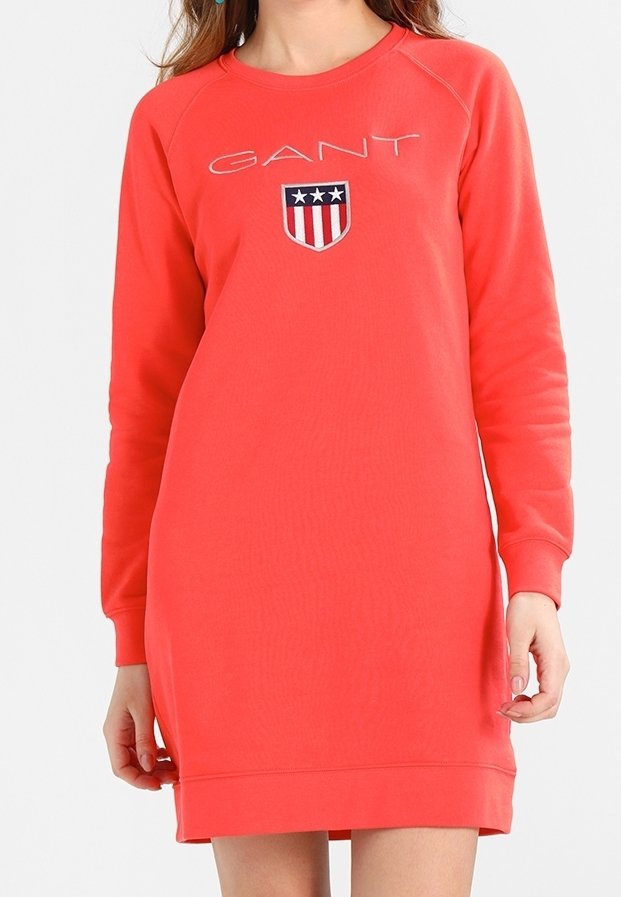 Femme portant une robe sweat-shirt corail à manches longues avec le logo GANT et un emblème en forme de bouclier avec des étoiles et des rayures sur la poitrine.
