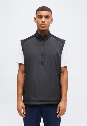 Homme portant un gilet noir sans manches avec fermeture éclair sur une chemise blanche à manches courtes, debout et regardant devant lui sur un fond uni.