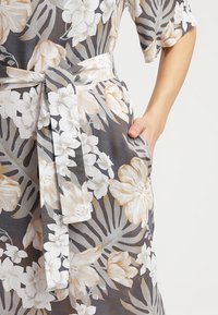 Robe fleurie grise avec des fleurs blanches et beiges, dotée d'une taille ceinturée, de manches courtes et de poches latérales, fabriquée en tissu léger.