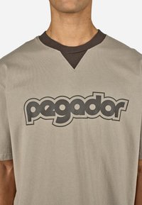 Beżowa koszulka z krótkimi rękawami, z ciemnobrązowym dekoltem w kształcie litery V i wyraźnym czarnym logo "pegador" z przodu. Miękka bawełniana tkanina.