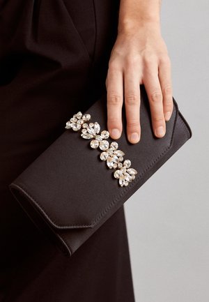 Hånd holder sort clutch taske med en række store, klare krystalblomsterdekorationer på forklappen.