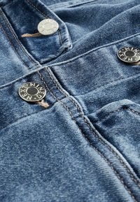 Tela de mezclilla que muestra tonalidades azules, con detalles visibles de costura y botones plateados brillantes con marca. Material texturizado y resistente.