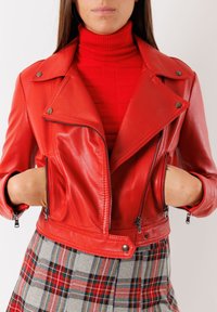 Giacca cropped in pelle rossa con dettagli in zip argentati, bottoni a scatto e colletto a pressione, abbinata a un dolcevita rosso e una gonna a scacchi.