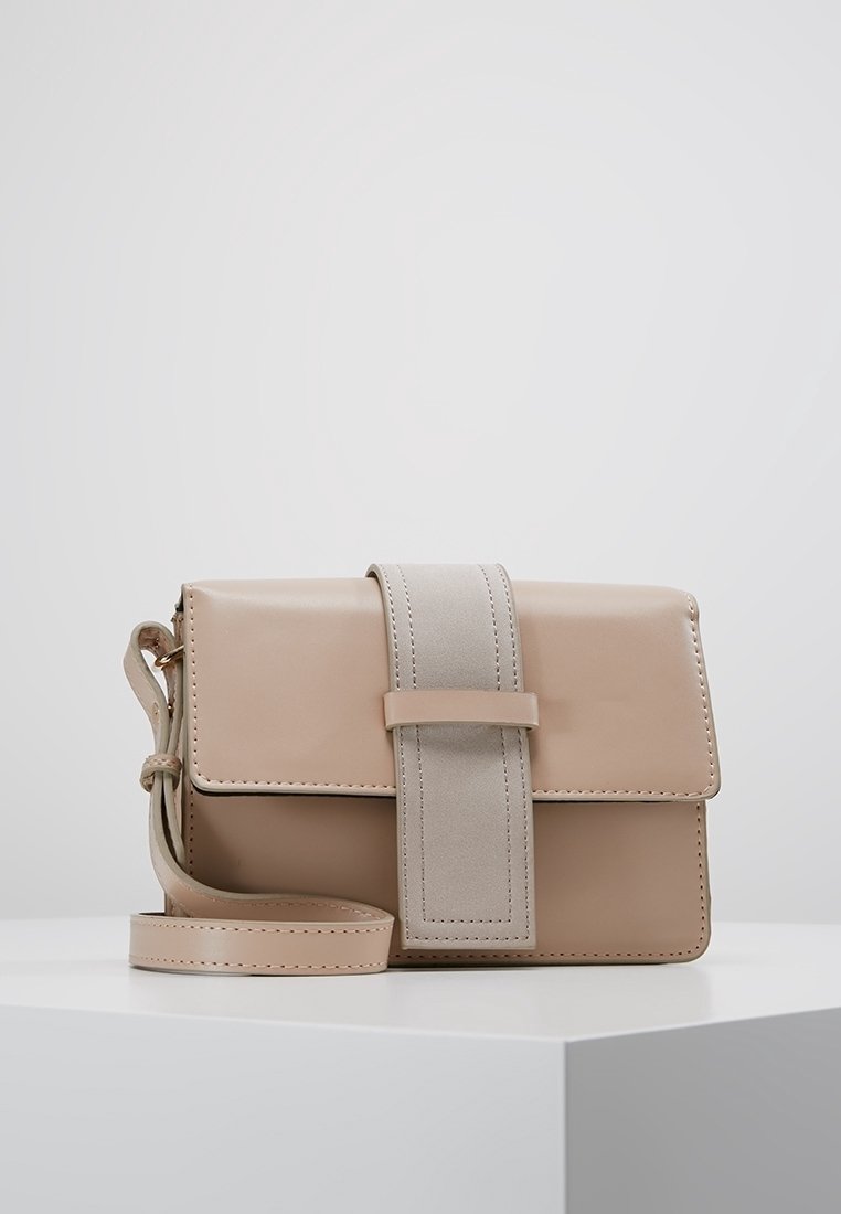 Béžová kožená crossbody taška se širokým semišovým popruhem a magnetickým uzávěrem, která leží na bílé podstavě na světlém šedém pozadí.