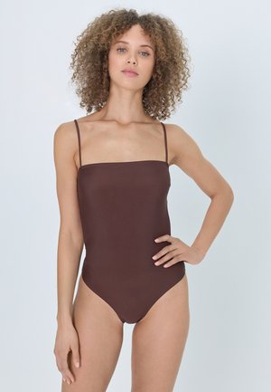 Frau mit lockigem Haar, die einen braunen, eng anliegenden ärmellosen Body mit dünnen Trägern trägt und mit einer Hand in der Hüfte steht.