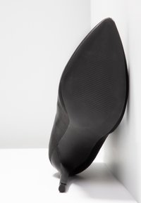 Chaussure noire à talon haut avec un bout pointu et un talon fin, présentant une texture lisse et une semelle rainurée, positionnée en angle.