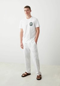 Camiseta blanca de algodón con un logo circular verde, combinada con pantalones cargo blancos y sandalias marrones. Diseño simple y material ligero.