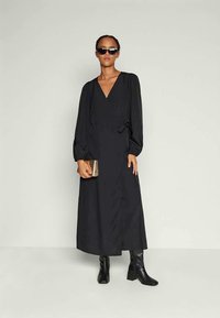 Mulher vestindo um longo vestido preto com mangas bufantes, botas pretas de tornozelo, óculos de sol em estilo gato e segurando uma pequena bolsa metálica.