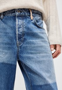 Hellblaue Jeans aus Denim mit ausgewaschener Textur, ausgestattet mit hohem Bund, vorderen Taschen und sichtbaren Nähdetails. Akzente in verschiedenen Farbtönen.