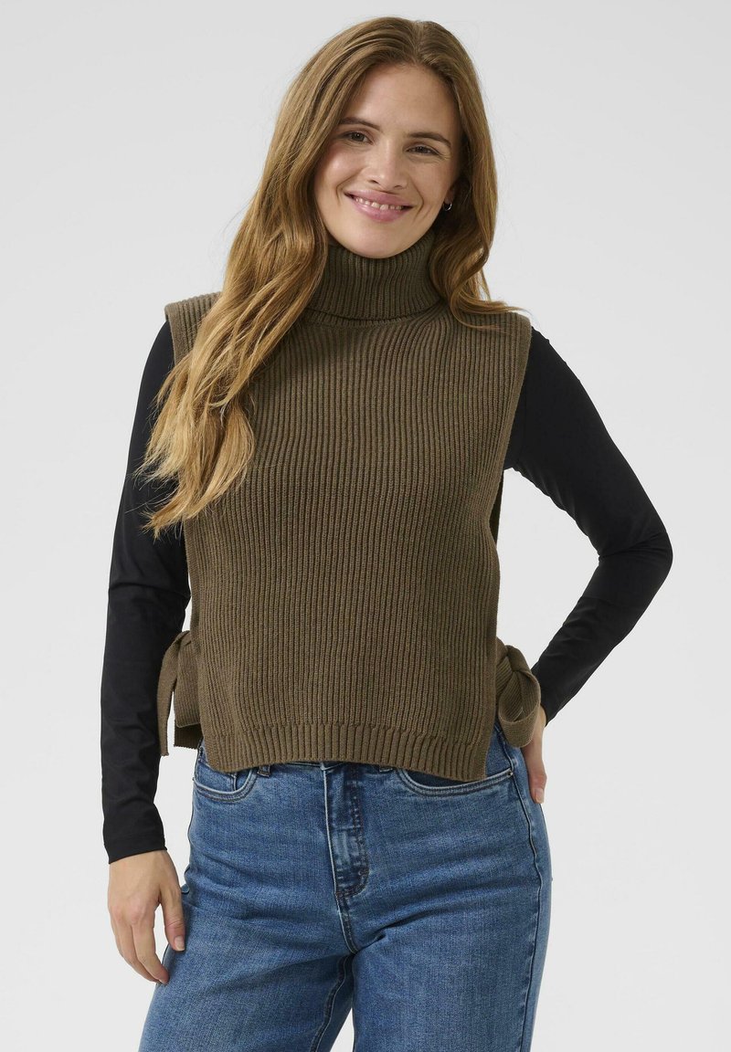 Débardeur en tricot vert olive à col roulé, avec une texture côtelée, des liens sur les côtés, porté sur une chemise à manches longues noire avec un jean bleu.