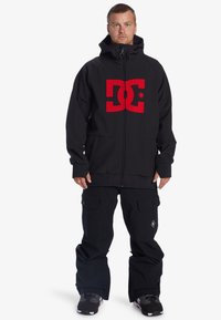 Čierna bunda na zips s veľkým červeným logom DC. V kombinácii s voľnými čiernymi nohavicami na sneh s bočnými vreckami. Štruktúrovaná tkanina.