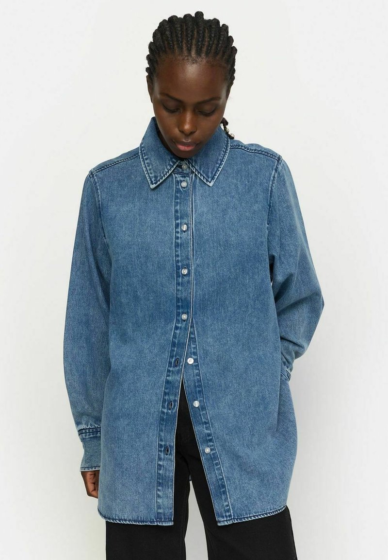 Džinsinė marškiniai su smailiu apykakle, mygtukų uždarymu ir ilgomis rankovėmis. Šviesiai mėlyna spalva, šiek tiek per didelis modelis, siuvimo detalės palei siūles.