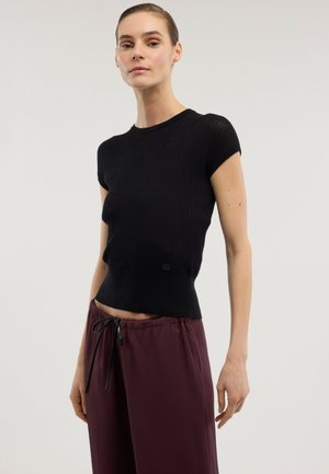 Kobieta w dopasowanym czarnym topie z krótkim rękawem i luźnych bordowych spodniach ze sznurkiem, stojąca na jasnym, jednolitym tle.