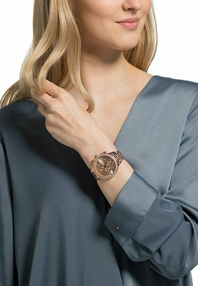 Orologio oro rosa con quadrante rotondo e caratteristiche di cronografo, montato su un bracciale metallico, indossato su una mano contro una blusa di satin blu.