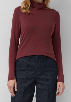 Kobieta w dopasowanym bordowym swetrze z golfem i granatowych, wysokich spodniach na szarym, jednolitym tle.