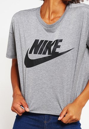 Person iført en grå Nike t-shirt med et stort sort swoosh-logo, som holder i kanten af trøjen med begge hænder.