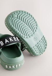 Groene rubberen instapsandalen hebben een gestructureerde zool met een visgraatpatroon en een zwarte band met witte letters.