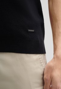 Maglione nero a maglia con tessuto texturizzato e orlo a coste. Etichetta metallica con "JOOP!" sul lato sinistro. Abbinato a pantaloni kaki, mostrando un dettaglio della mano.