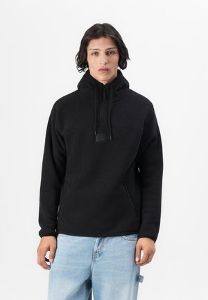 Pullover in pile nero con collo alto, zip frontale e tasche laterali. Texture morbida e design minimalista. Modello che indossa jeans blu larghi.