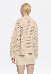 Maglione beige a maglia grossa con un design a rete aperta, polsini e orlo a coste. Indossato sopra una gonna beige aderente, che mette in mostra contrasti di texture.