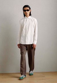 Witte button-up shirt, bruine broek en groen gestreepte sneakers. Zwarte zonnebril maakt de look compleet. Eenvoudig ontwerp, zachte textuur.