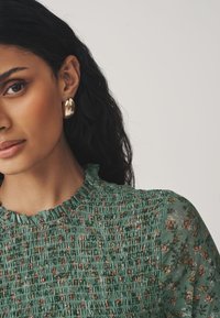 Green, textured blouse with a smocked design and floral patterns. Features a ruffled collar and a woman wearing gold, oval-shaped earrings.