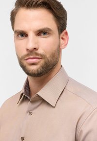 Hellbraune Button-Up-Bluse aus strukturiertem Stoff mit einem gemusterten Kragen-Innenfutter. Braune Knöpfe und ein klassisches Kragen-Design.