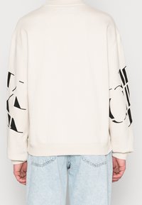 Personne portant un sweat crème avec un grand texte graphique noir sur les manches et un jean en denim bleu clair, vu de dos.