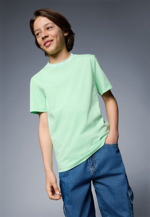 T-shirt en coton vert menthe à manches courtes, avec un col rond et une bande logo blanche au niveau du col, associé à un short cargo en denim bleu.