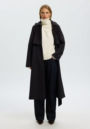 Femme portant un long manteau noir, un pull à col roulé blanc, un pantalon large foncé et des chaussures pointues marron sur un fond clair uni.