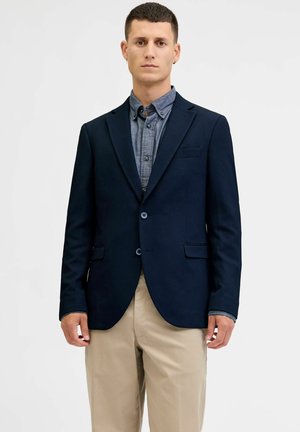 Mann trägt einen marineblauen Blazer über einem blauen Hemd mit Knöpfen und beigen Hosen und steht vor einem einfarbigen hellen Hintergrund.