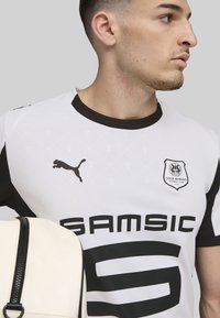Camiseta de fútbol blanca con acentos negros, que presenta el logo de Puma, el escudo del Stade Rennais y grandes letras "SAMSIC". Textura de tela suave.