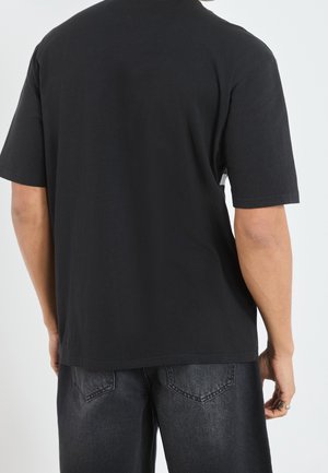 Persoon draagt een effen zwart T-shirt met korte mouwen en versleten zwarte spijkerbroek, staand met de rug naar voren tegen een lichtgrijze achtergrond.