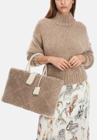 Beige quilted faux fur handbag with white leather accents and detachable tag, carried with a ribbed brown turtleneck sweater.