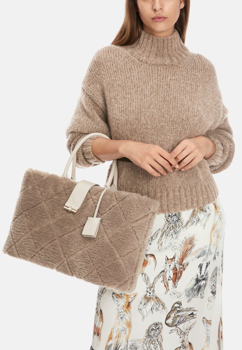 Beige quilted faux fur handbag with white leather accents and detachable tag, carried with a ribbed brown turtleneck sweater.