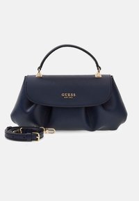 Μπλε σκούρα τσάντα Guess με χρυσά μεταλλικά μέρη, επάνω λαβή, λογότυπο μπροστά και αποσπώμενο λουρί ώμου τυλιγμένο δίπλα στην τσάντα σε ανοιχτό φόντο.