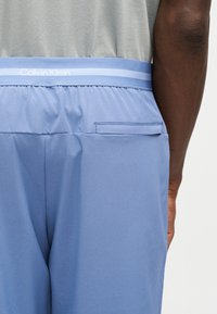 Κοντινό πλάνο ατόμου που φοράει ανοιχτό μπλε παντελόνια Calvin Klein με ελαστική μέση και πίσω τσέπη, συνδυασμένα με γκρι πουκάμισο.