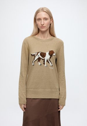 Femme aux longs cheveux blonds portant un pull beige avec un motif de chien marron et blanc et une jupe marron sur un fond clair uni.
